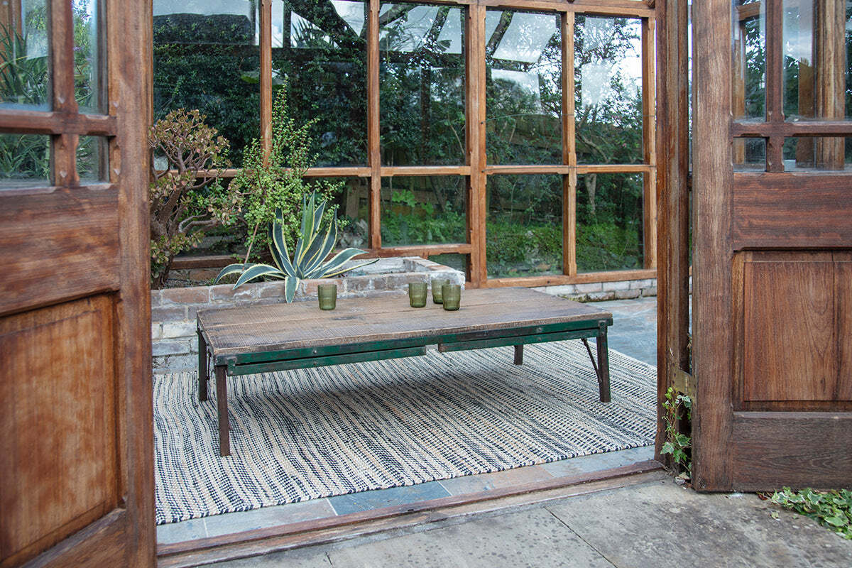 The Ishan Reclaimed Folding Dining & Coffee Table by nkuku is set with three green glasses on a striped rug. Placed before large wood-framed windows, it opens to a cozy indoor-outdoor area with green plants and wooden doors.