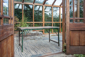 The nkuku Ishan Reclaimed Folding Dining & Coffee Table rests on a woven rug in a sunlit greenhouse, with open wooden doors framing vibrant greenery outside.