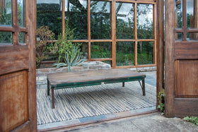 The Ishan Reclaimed Folding Dining & Coffee Table by nkuku, featuring a green frame, stands on a striped rug near wide wooden-framed glass doors with views of plants, bricks, and large windows reflecting garden scenery.