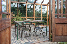 The nkuku Ishan Reclaimed Folding Dining & Coffee Table is styled with green chairs in a greenhouse. Plates and glasses rest atop the reclaimed wood, as sunlight through large windows creates a warm, natural ambience.