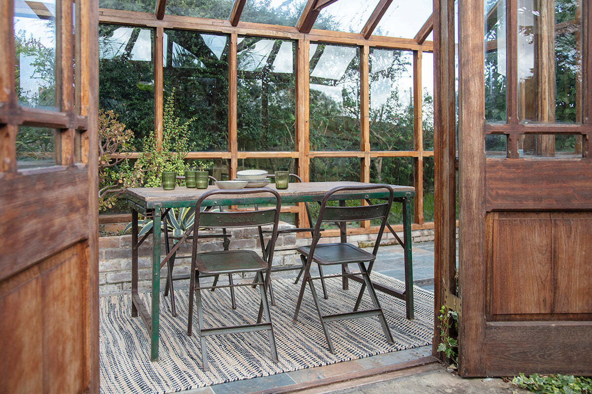 The nkuku Ishan Reclaimed Folding Dining & Coffee Table is styled with green chairs in a greenhouse. Plates and glasses rest atop the reclaimed wood, as sunlight through large windows creates a warm, natural ambience.