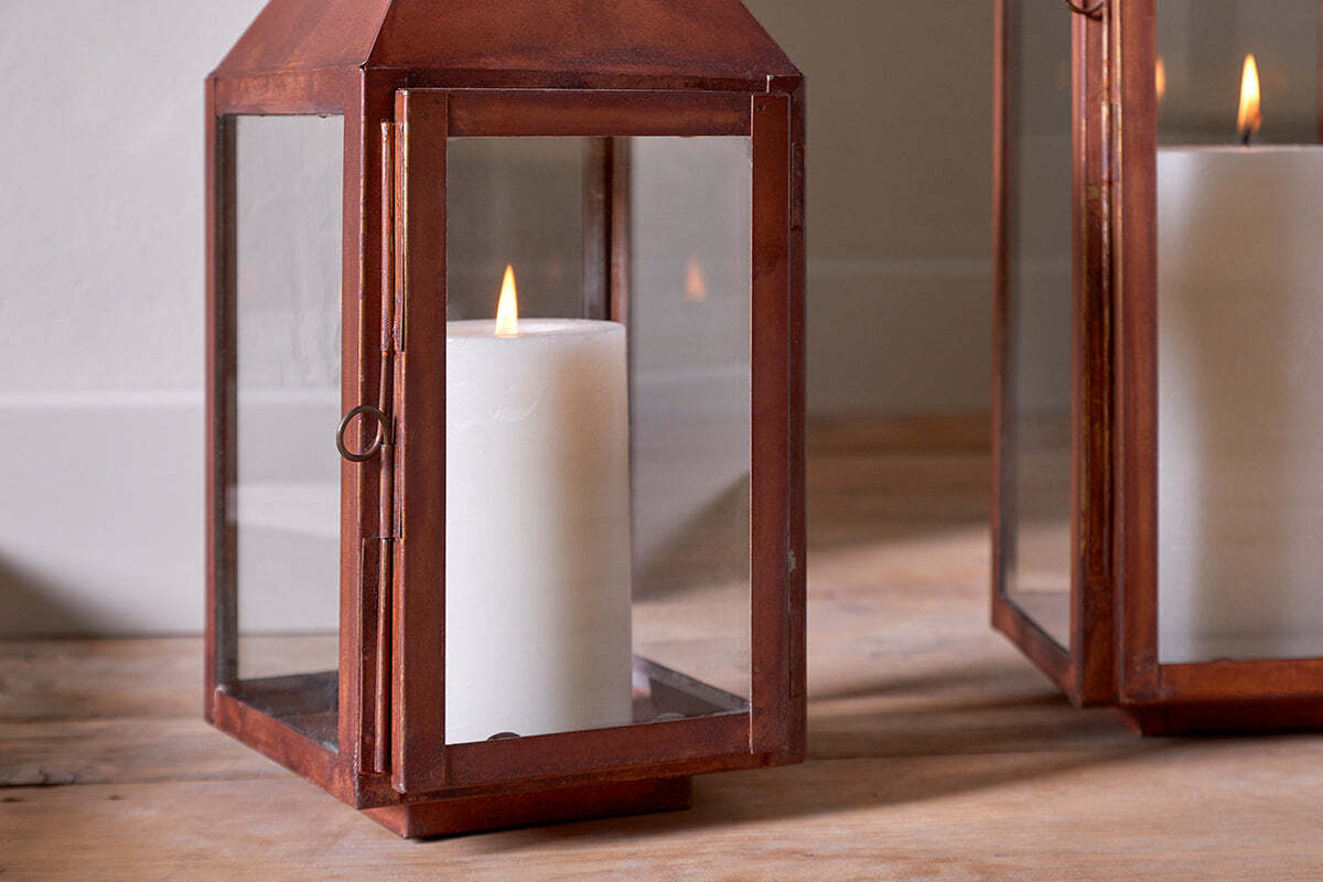 A lit white pillar candle glows inside the nkuku Isko Lantern - Rust, featuring a rectangular glass and copper design with a rustic finish on a wooden floor—perfect for adding cozy accents to your garden seating.