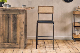 The Iswa Leather & Cane Counter Chair - Aged Black by nkuku features a woven cane back, black metal frame, and cushioned leather seat. It sits on a wooden floor near a rustic bar with minimal decor, with shelving visible in the background.