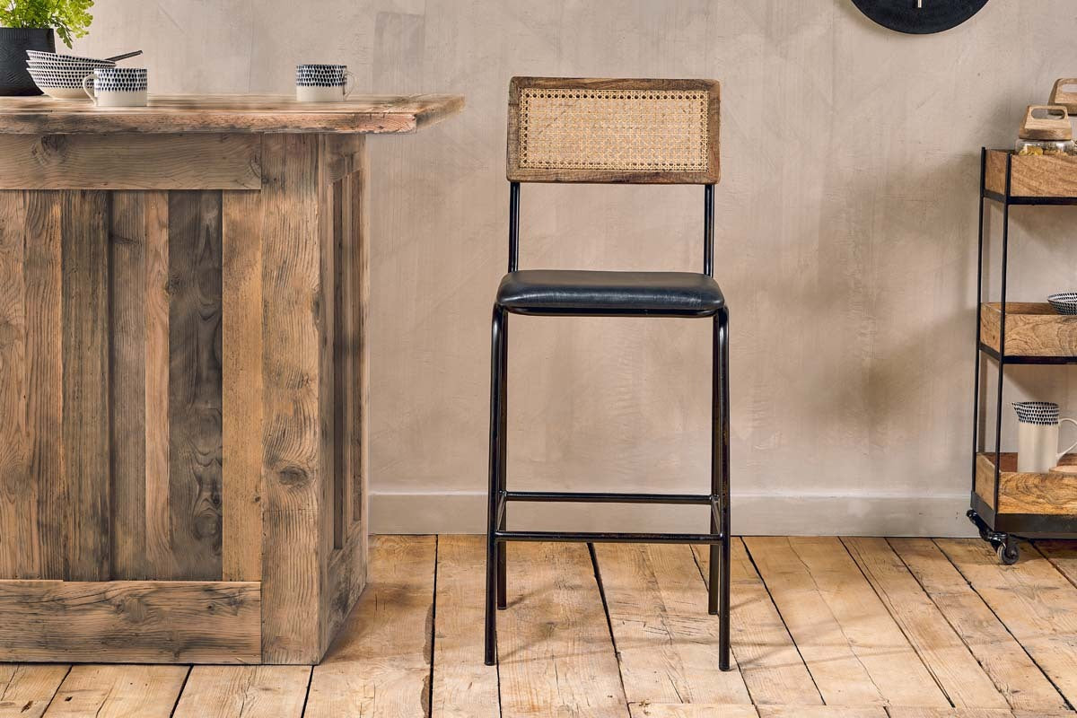 The Iswa Leather & Cane Counter Chair - Aged Black by nkuku features a woven cane back, black metal frame, and cushioned leather seat. It sits on a wooden floor near a rustic bar with minimal decor, with shelving visible in the background.