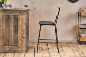 The Iswa Leather & Cane Counter Chair by nkuku, featuring an aged black frame, black leather seat, and light cane backrest, is placed by a rustic wooden bar on wooden flooring with a small cart against a beige wall in the background.