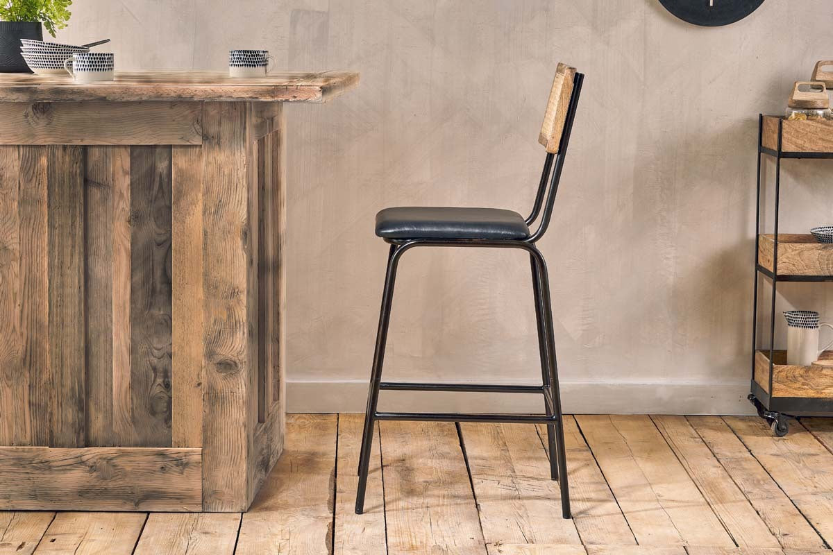 The Iswa Leather & Cane Counter Chair by nkuku, featuring an aged black frame, black leather seat, and light cane backrest, is placed by a rustic wooden bar on wooden flooring with a small cart against a beige wall in the background.