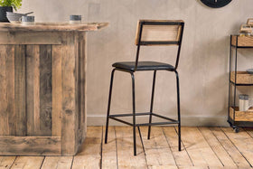 The Iswa Leather & Cane Counter Chair by nkuku, featuring a woven cane back, black metal frame, and cushioned black leather seat in aged black, stands on a rustic wooden floor beside a wooden bar counter in a cozy, modern room.