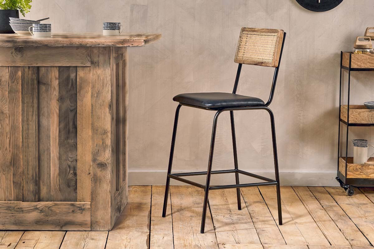 A rustic kitchen scene with the nkuku Iswa Leather & Cane Counter Chair in Aged Black, featuring a black metal frame, woven cane back, and black leather seat beside a weathered wood bar on wooden floors with decor and a rolling cart in the background.