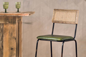 The Iswa Leather & Cane Counter Chair by nkuku features a woven cane back, black metal legs, and a rich green leather seat—ideal for stylish kitchen island seating beside rustic wooden bars and green glass goblets against beige walls.