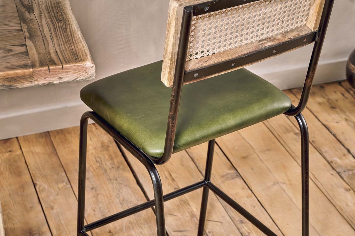 The Iswa Leather & Cane Counter Chair by nkuku features a rich green cushioned seat, metal frame, and woven rattan cane back—perfect for kitchen island seating against a beige wall and rustic table on a wooden floor.