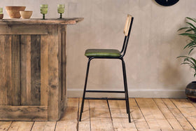 A rustic wooden bar sits on a light wood floor next to the nkuku Iswa Leather & Cane Counter Chair in rich green. Three green glasses, wooden bowls, and a leafy potted plant decorate the space against the beige wall.