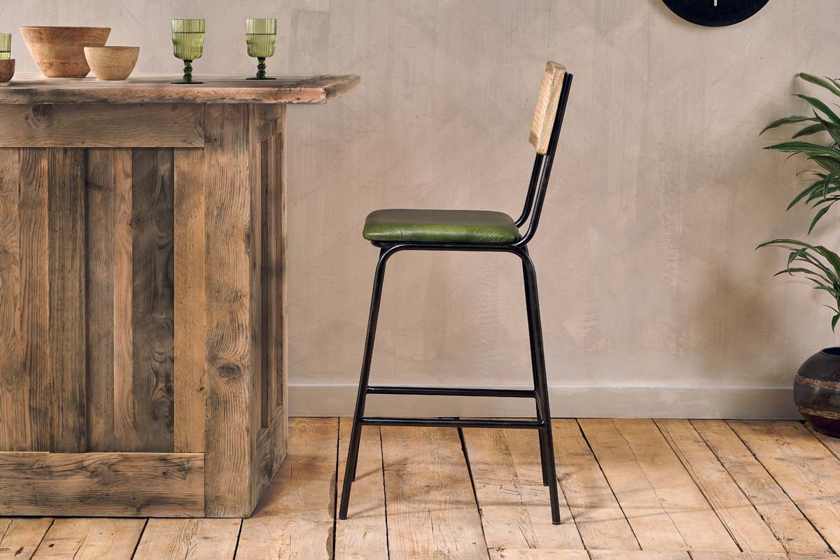 A rustic wooden bar sits on a light wood floor next to the nkuku Iswa Leather & Cane Counter Chair in rich green. Three green glasses, wooden bowls, and a leafy potted plant decorate the space against the beige wall.