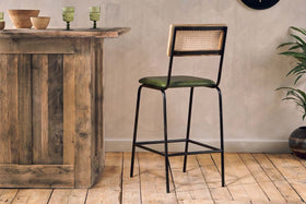 The Iswa Leather & Cane Counter Chair by nkuku features a rich green cushioned seat, black metal frame, and light wood cane backrest—perfect as kitchen island seating. Green glassware and bowls are on the bar, with a plant to the right.