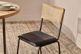 The Iswa Leather & Cane Dining Chair - Black by nkuku, featuring a black goat leather seat and woven backrest, rests on a rustic mango wood floor beside a round wooden table set with bowls and chopsticks.