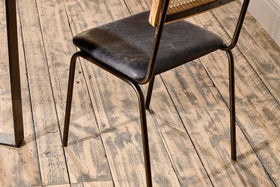 Close-up of the Iswa Leather & Cane Dining Chair - Black by nkuku, crafted with black goat leather and a mango wood frame, shown on distressed wooden floorboards.