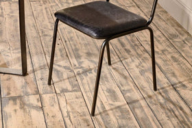 The nkuku Iswa Leather & Cane Dining Chair in black, featuring a slim metal frame and gently aged goat leather seat, is set on a distressed light wood floor with visible grain. A metal table leg appears on the image's left side.