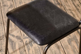 Close-up of the Iswa Leather & Cane Dining Chair by nkuku in black. The textured goat leather seat features visible grain and scuff marks, adding character against a distressed wooden floor.