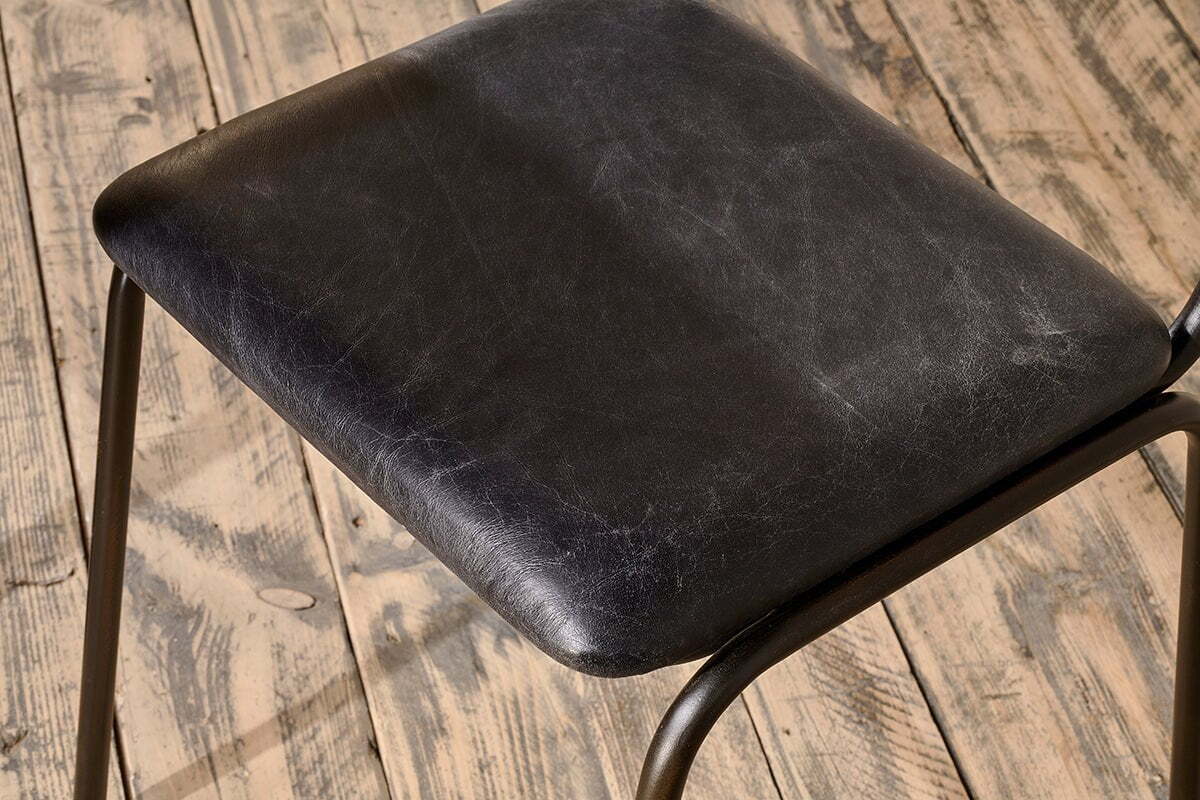 Close-up of the Iswa Leather & Cane Dining Chair by nkuku in black. The textured goat leather seat features visible grain and scuff marks, adding character against a distressed wooden floor.