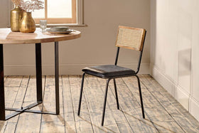 The Iswa Leather & Cane Dining Chair - Black by nkuku showcases a mango wood frame, black goat leather seat, and cane back, positioned beside a round wooden dining table with black metal legs on a rustic wooden floor.