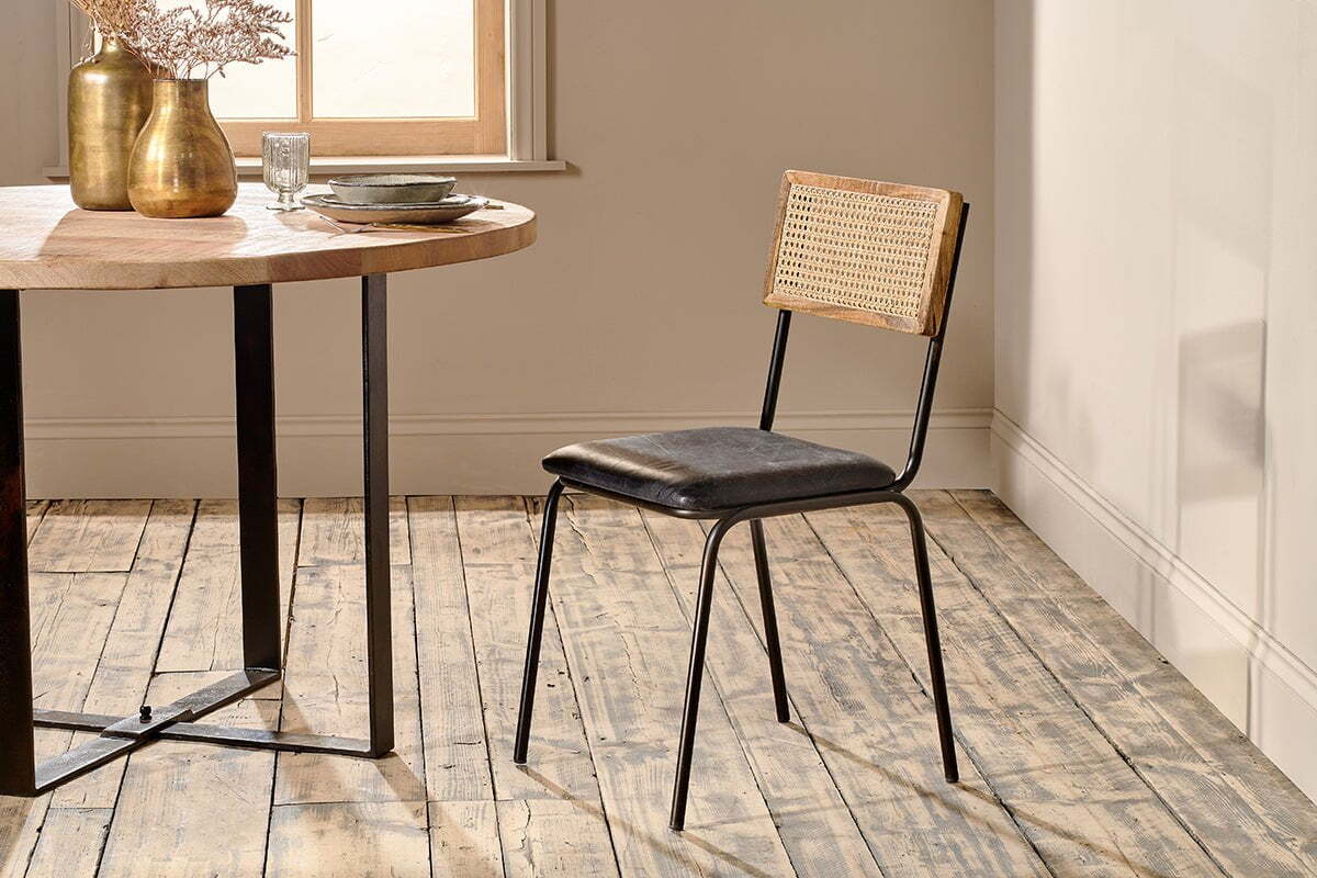 The Iswa Leather & Cane Dining Chair - Black by nkuku showcases a mango wood frame, black goat leather seat, and cane back, positioned beside a round wooden dining table with black metal legs on a rustic wooden floor.