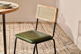 The nkuku Iswa Leather & Cane Dining Chair - Green, featuring an industrial black iron frame and light wood backrest, is placed on a rustic floor beside a round table. The room has beige walls and white baseboards.