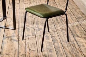 The Iswa Leather & Cane Dining Chair - Green by nkuku, featuring a green cushioned seat and black metal legs, is placed on a rustic wooden floor near an off-white wall and next to a black metal table.