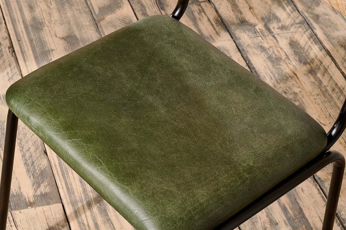 Close-up of the Iswa Leather & Cane Dining Chair in green by nkuku, featuring dark green goat leather with subtle creases, a black iron frame, and a cane seat, set on a rustic wooden floor.
