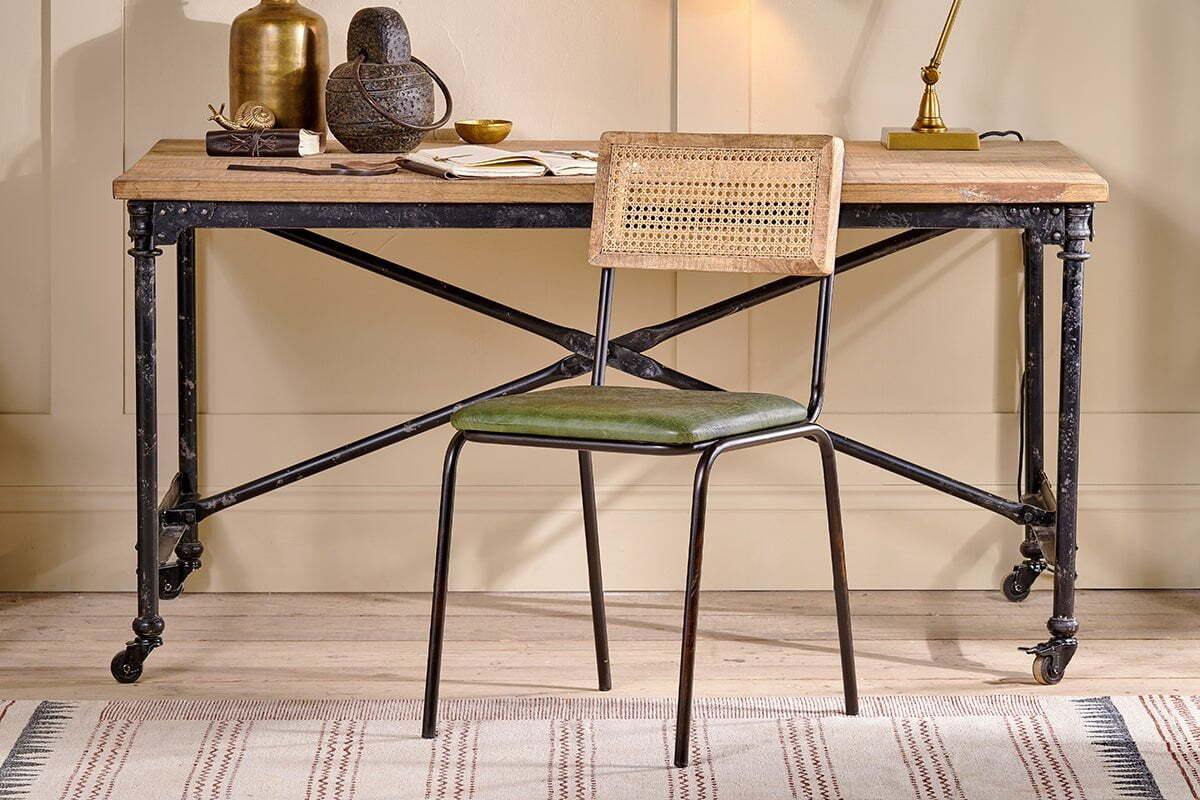A rustic wooden desk with an industrial black iron frame and wheels is paired with the nkuku Iswa Leather & Cane Dining Chair in green. Decorative items sit atop the desk.