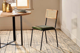 The rustic dining set features a round wooden table with an industrial black iron frame, paired with the nkuku Iswa Leather & Cane Dining Chair in green, which has a cane-backed wooden frame and a green upholstered seat.