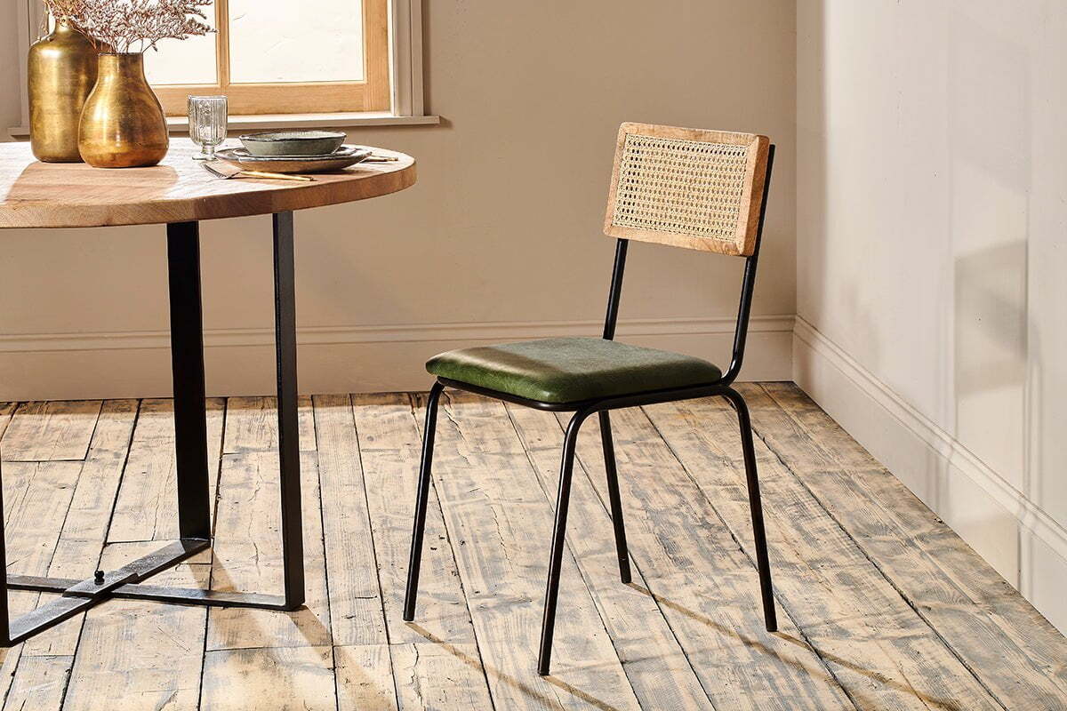 The rustic dining set features a round wooden table with an industrial black iron frame, paired with the nkuku Iswa Leather & Cane Dining Chair in green, which has a cane-backed wooden frame and a green upholstered seat.