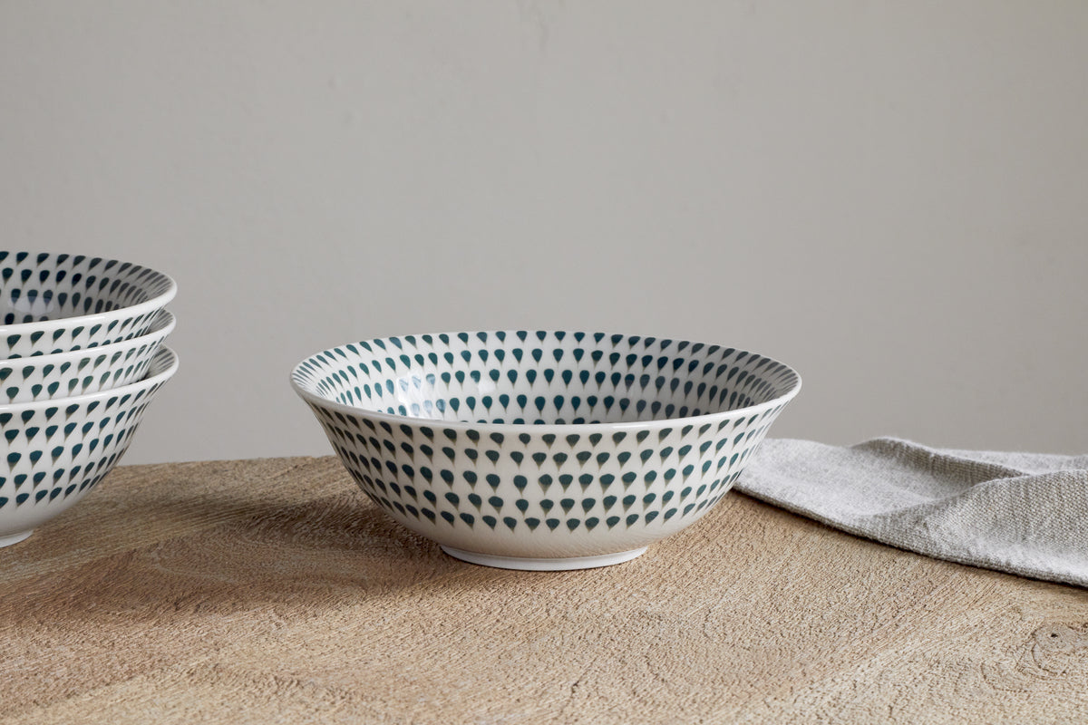 A Juniper Drop Dinnerware Set - Cream & Juniper (Set of 12) by nkuku features a white ceramic bowl with a blue drop pattern on a wooden surface, beside a folded light gray cloth and more stacked bowls in the background.