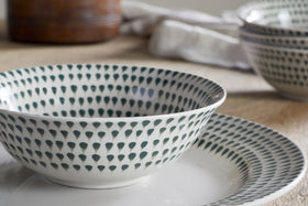 A close-up of the nkuku Juniper Drop Cereal Bowl - Cream & Juniper, with its green drop pattern, sits on a matching plate. In the background, a similar bowl and textured cloth complete this inviting dining scene.