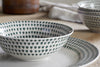 A close-up of the nkuku Juniper Drop Dinnerware Set - Cream & Juniper (Set of 12) shows a bowl and plate with a repeating green drop design atop a beige tablecloth, with another matching bowl and a blurred towel in the background.