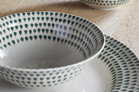 A close-up of the nkuku Juniper Drop Cereal Bowl in Cream & Juniper, featuring a green drop pattern on the rim, perfect for everyday dining, resting on a beige surface with another bowl partially visible in the background.