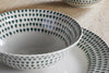 A close-up of the nkuku Juniper Drop Dinnerware Set - Cream & Juniper (Set of 12), showing a bowl and plate with evenly spaced juniper green drops on the rim, displayed on a light textured surface.