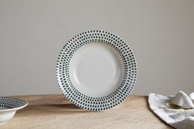 The Juniper Drop Pasta Bowl - Cream & Juniper by nkuku, a white ceramic bowl with small dark green drops around the rim, rests on a wooden surface with a cloth napkin and brass spoon partially visible to the right.