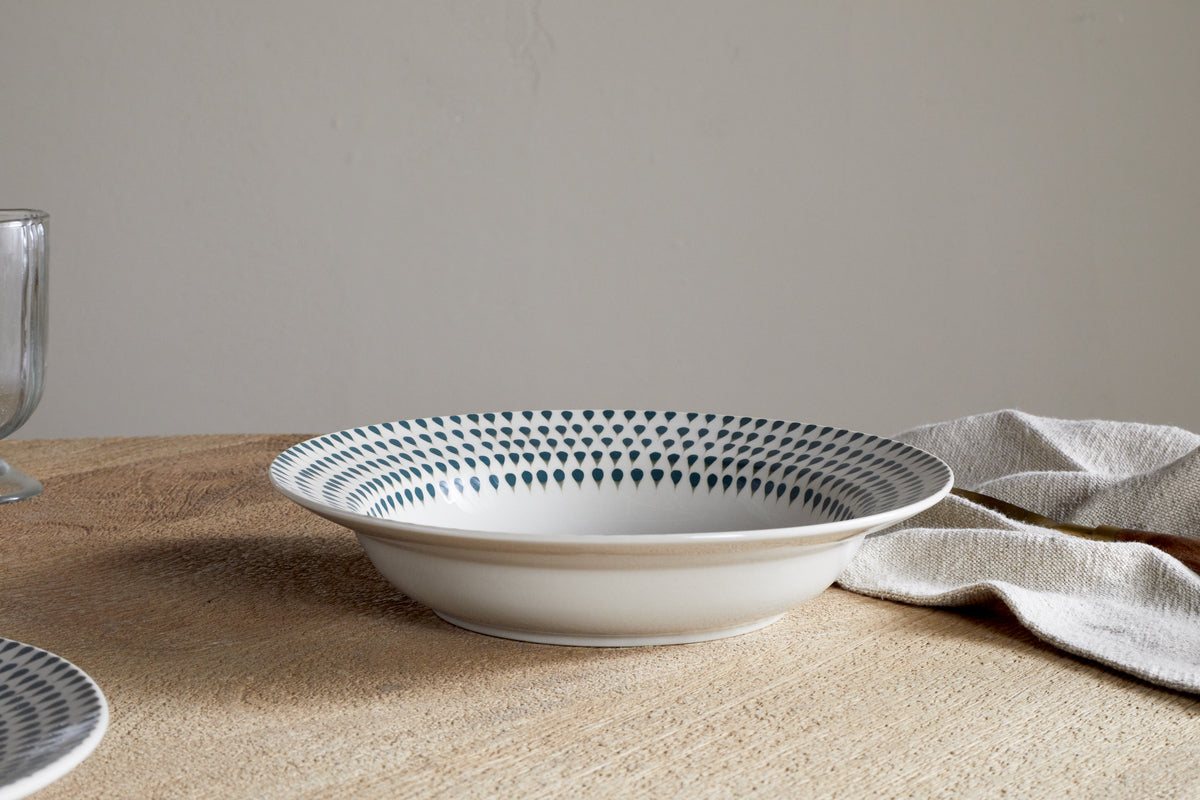 The Juniper Drop Pasta Bowl - Cream & Juniper by nkuku is a contemporary white ceramic bowl with a blue dotted rim, shown on a wooden table beside a beige napkin, glass, and partial plate.