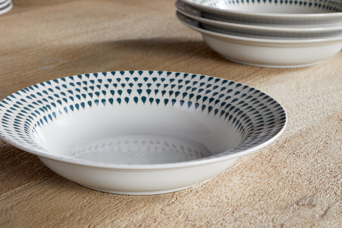 A close-up of the nkuku Juniper Drop Pasta Bowl in Cream & Juniper, featuring dark blue teardrop patterns around the rim, sits on a wooden surface with stacks of matching contemporary tableware in the background.