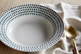 The Juniper Drop Pasta Bowl - Cream & Juniper by nkuku, featuring a blue teardrop rim design, sits on a beige cloth beside a gold spoon with wooden handle, highlighting contemporary tableware on a light wooden surface.