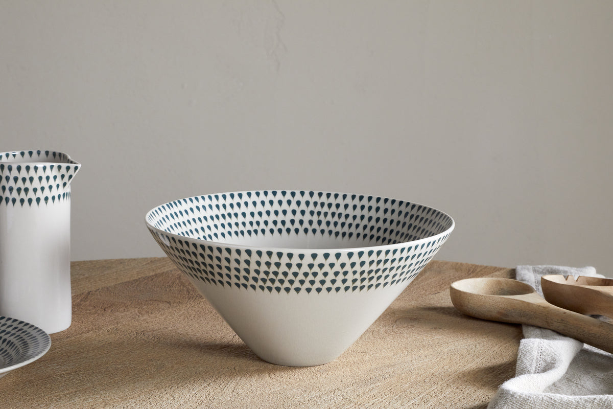 The nkuku Juniper Drop Serving Bowl in cream and juniper, featuring dark blue teardrop shapes along the wide rim, rests on a wooden surface beside matching dishes and a light cloth, creating an elegant centerpiece.