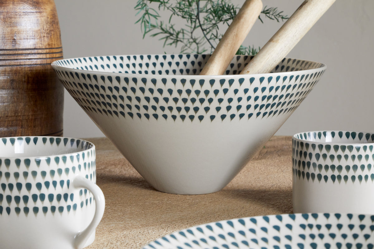 The nkuku Juniper Drop Serving Bowl - Cream & Juniper (Medium), featuring a blue teardrop pattern, holds two wooden utensils on a textured surface. Matching cups and plate, plus a plant and wooden jar, complete the inviting centerpiece.
