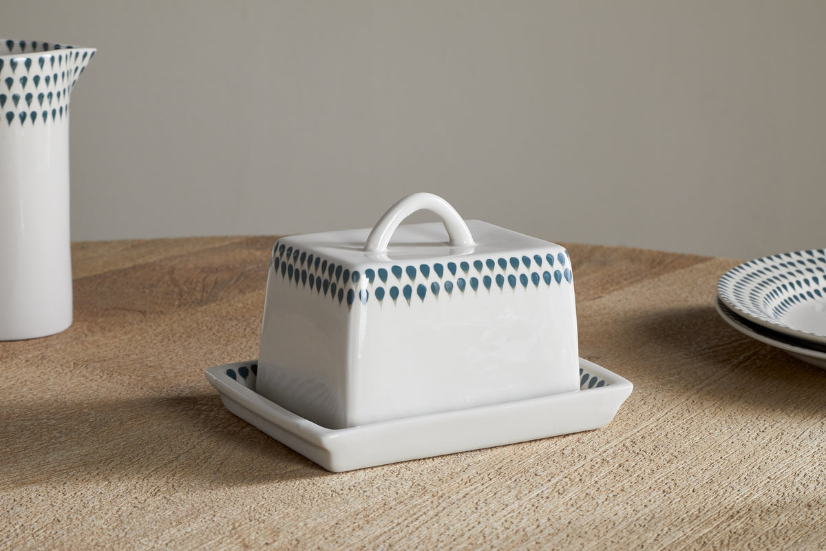The nkuku Juniper Drop Butter Dish - Cream & Juniper features a white ceramic base and lid with a blue teardrop pattern, displayed on a light wooden table next to matching plates.