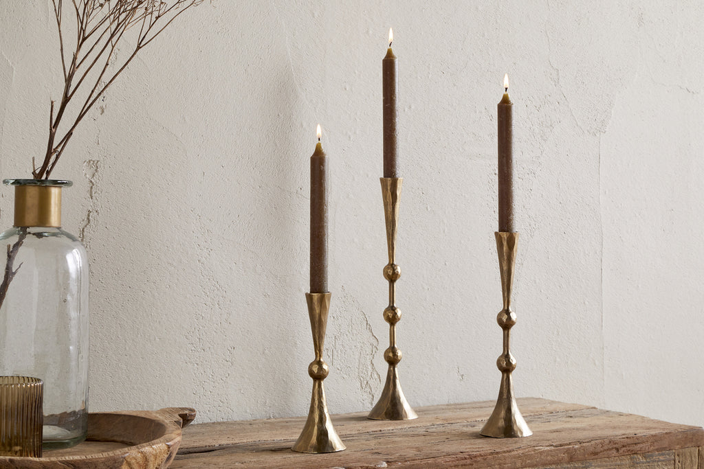 Three tall brown candles in nkuku’s Javanee Candle Holders - Antique Brass are lit and arranged on a rustic wooden table beside a clear glass vase with dried branches and a small ribbed glass container, all set against a textured white wall.