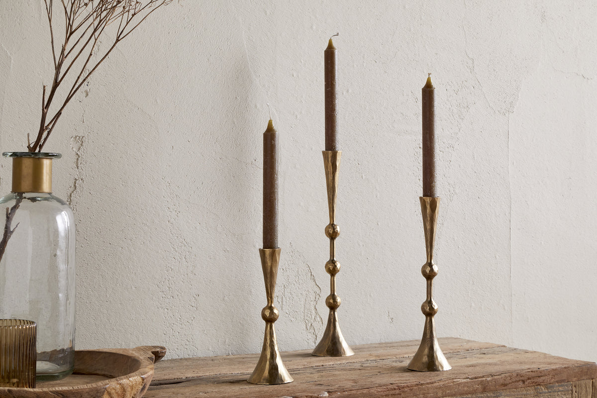 Three nkuku Javanee Candle Holders in antique brass with brown taper candles are arranged on a rustic wooden surface. Nearby, a clear glass vase of dried branches adds to the sculptural scene against a textured light wall.