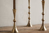 Three nkuku Javanee Candle Holders in antique brass, featuring a hammered texture, geometric shapes, and spherical accents, stand on a rustic wooden surface. No candles are inserted.