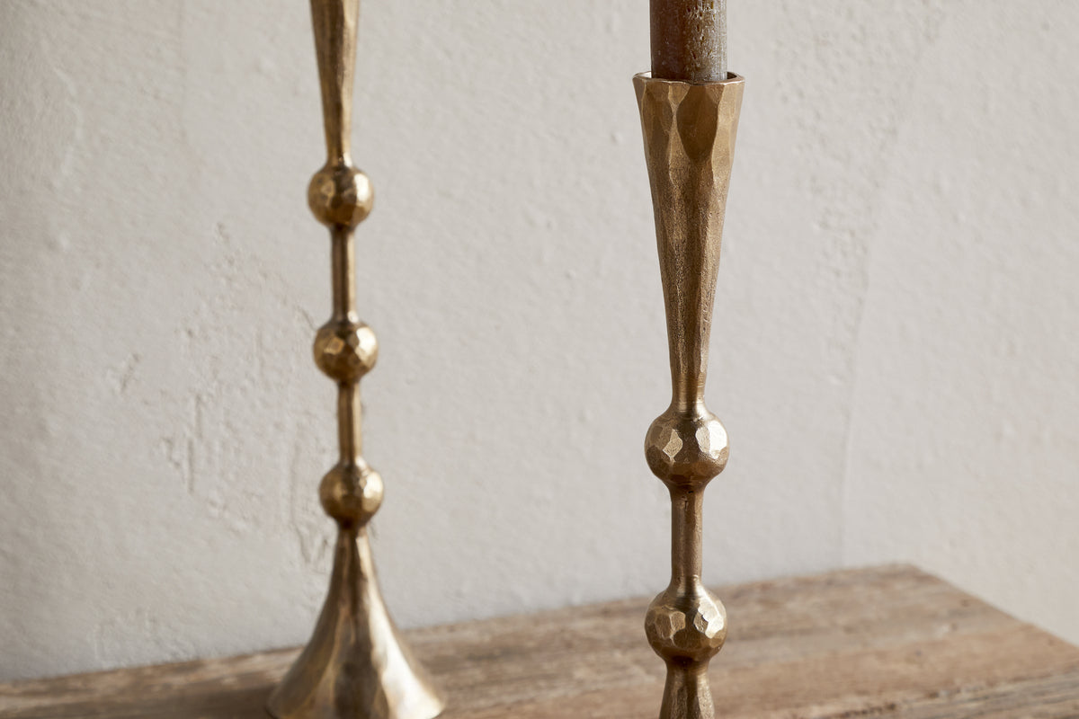 Two nkuku Javanee Candle Holders in antique brass, featuring a geometric hammered texture and spherical accents, stand on a rustic wooden surface with one partially visible on the left against a white textured wall.
