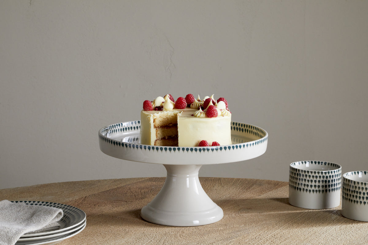 A small frosted cake with raspberries and cream sits on the nkuku Juniper Drop Ceramic Stand - Cream & Juniper, placed on a wooden table beside green drop-patterned cups and a folded napkin. One slice has been removed.