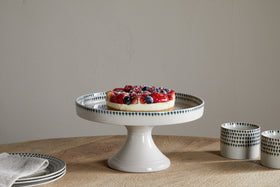 A fruit tart topped with strawberries and blueberries rests on the nkuku Juniper Drop Ceramic Stand - Cream & Juniper, set on a wooden table with a stack of plates, a folded napkin, and two patterned cups nearby.