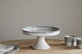The nkuku Juniper Drop Ceramic Stand - Cream & Juniper, featuring a green drop rim design, sits on a wooden table with matching plates, a napkin, and two small cups against a plain light background.
