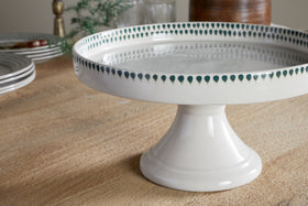 The nkuku Juniper Drop Ceramic Stand - Cream & Juniper features a green drop border and sits on a wooden table with stacked plates, glasses, and blurred decor in the background.